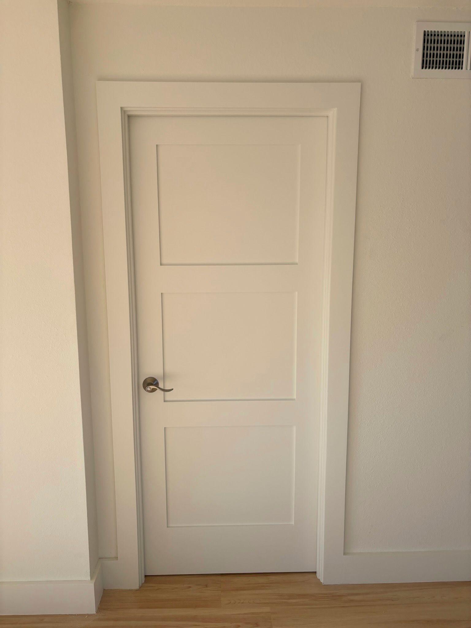 White three-panel interior door with silver handle and trim. Located between white walls and wooden floor.