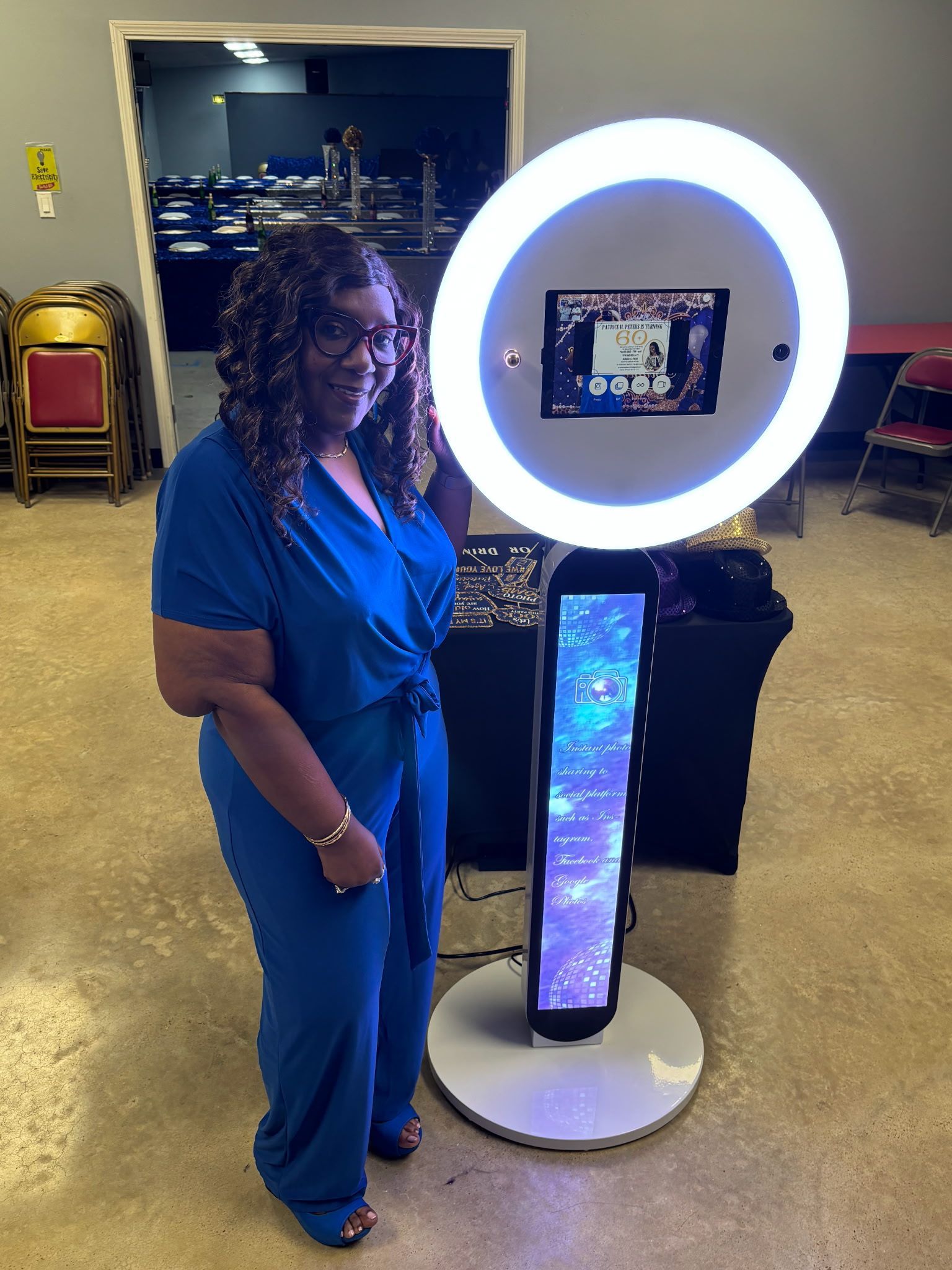Woman in blue jumpsuit poses beside a photo booth with a light ring; party setting.