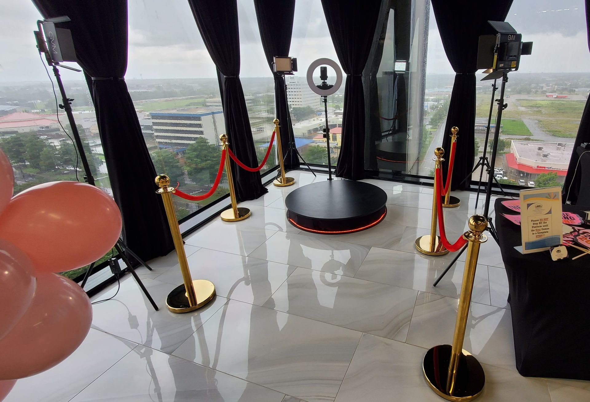 Photo booth setup with red ropes, black curtains, and a ring light in a venue with a city view.