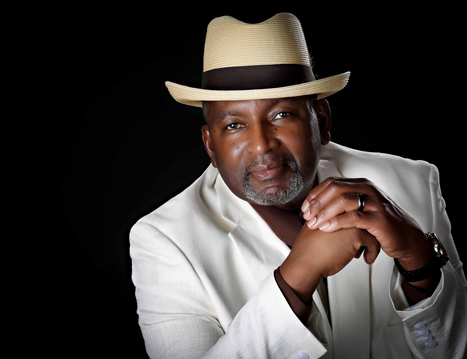 Man in white jacket and straw hat, hands clasped, smiling, against black backdrop.
