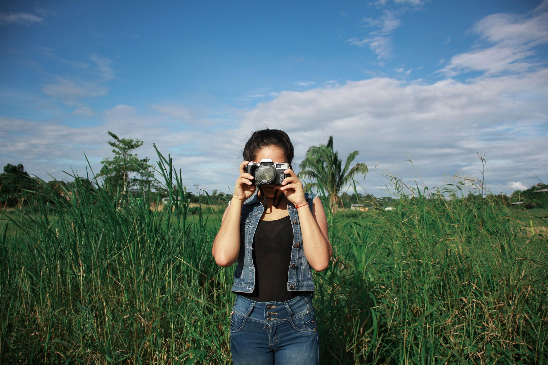 Woman with a camera taking a photo outdoors, surrounded by tall grass, under a blue sky.