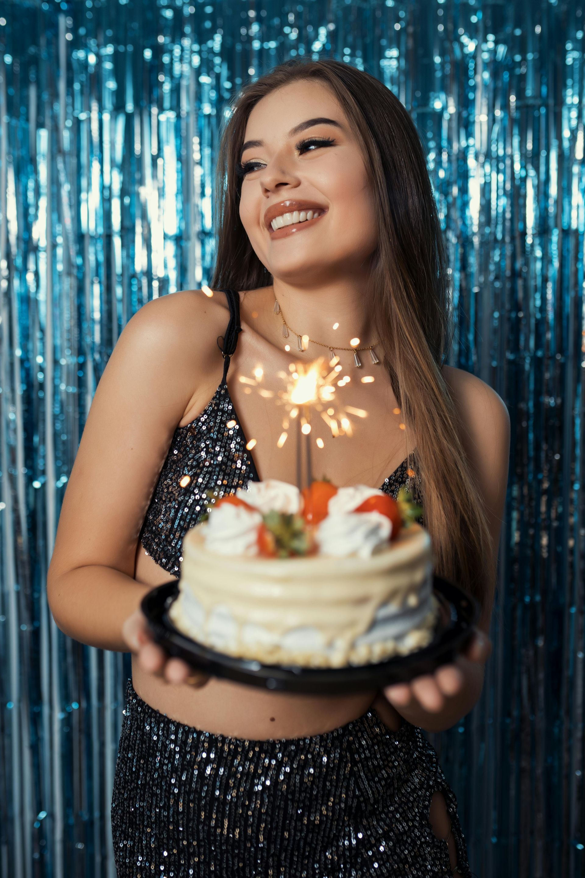 Woman holding a birthday cake with a sparkler, smiling, against a blue backdrop.