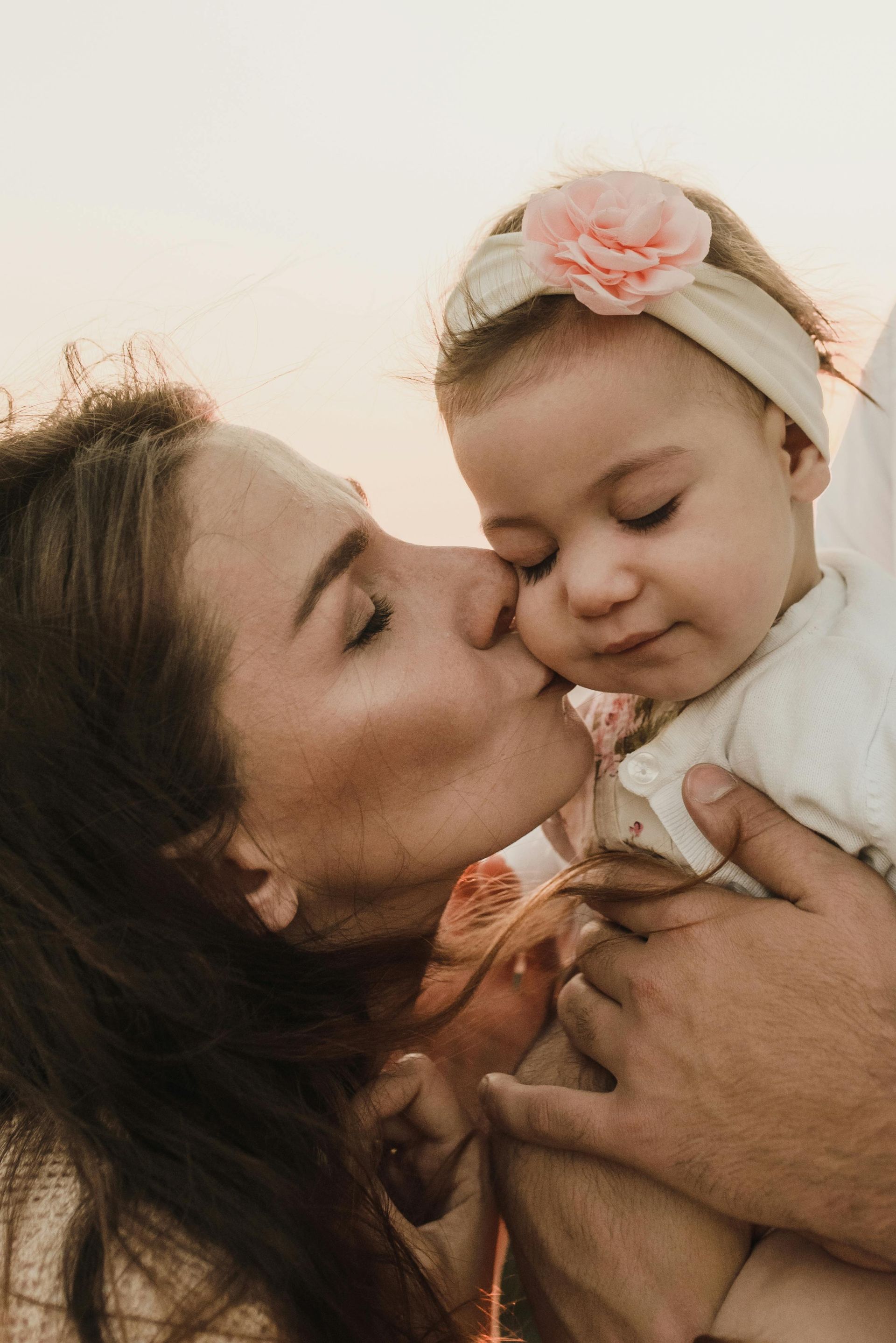 Mother kisses a baby's cheek, both illuminated in warm light. The baby wears a headband with a flower.