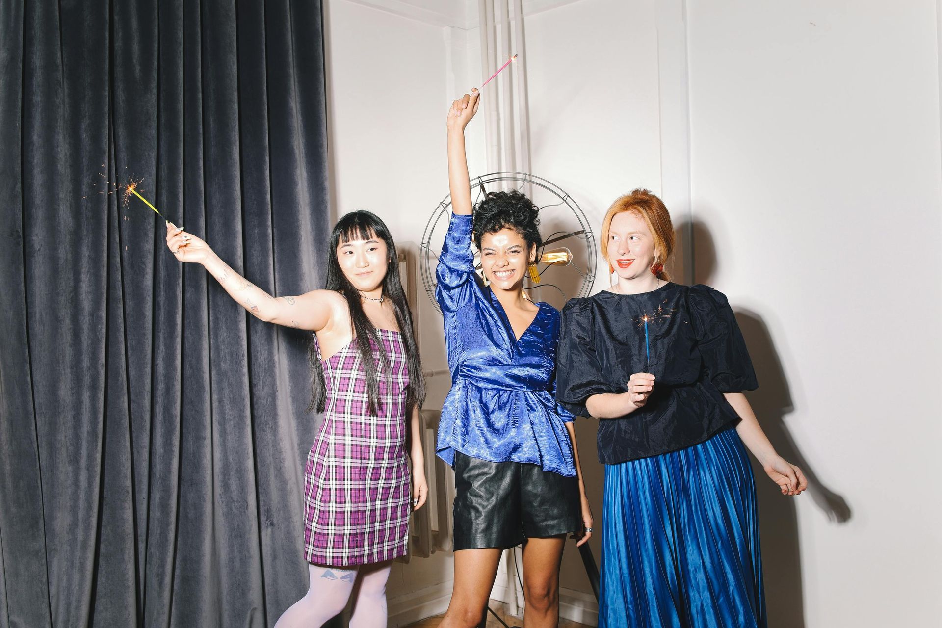 Three diverse women with sparklers celebrate in a room with a curtain and fan.