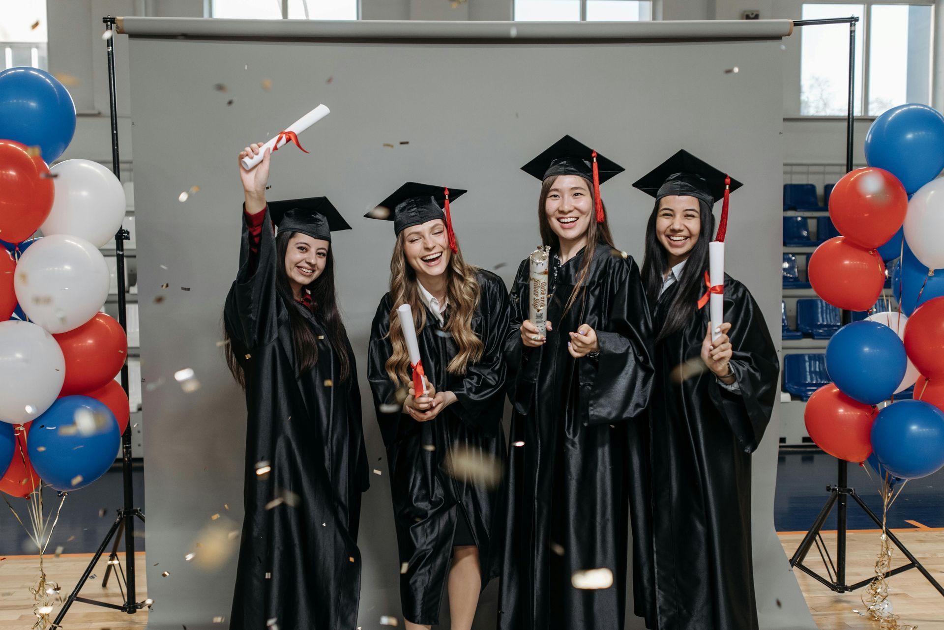 Four graduates in black gowns and mortarboards with diplomas, red, white, and blue balloons, and confetti.