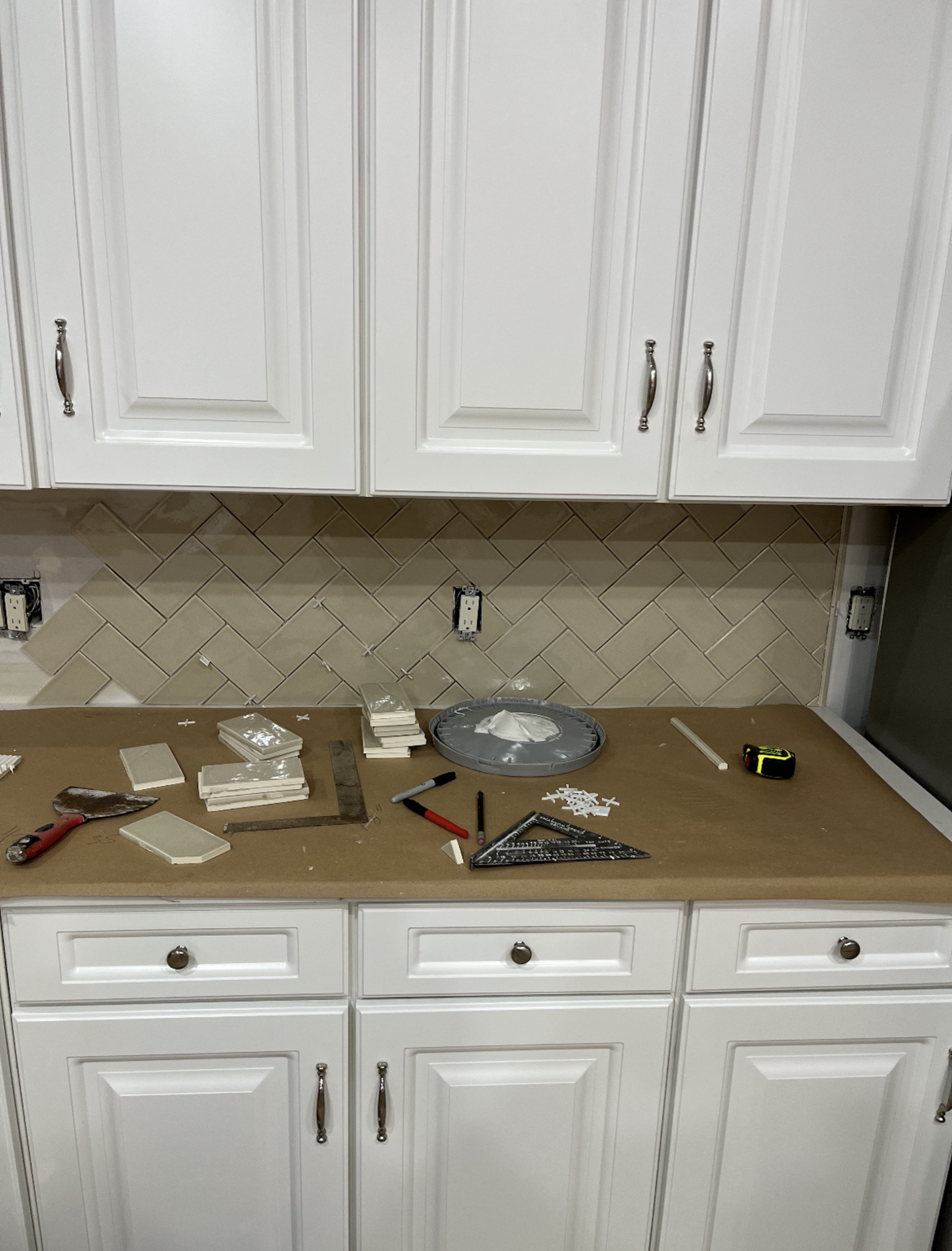 White kitchen cabinets with backsplash tile being installed; tools on the countertop.