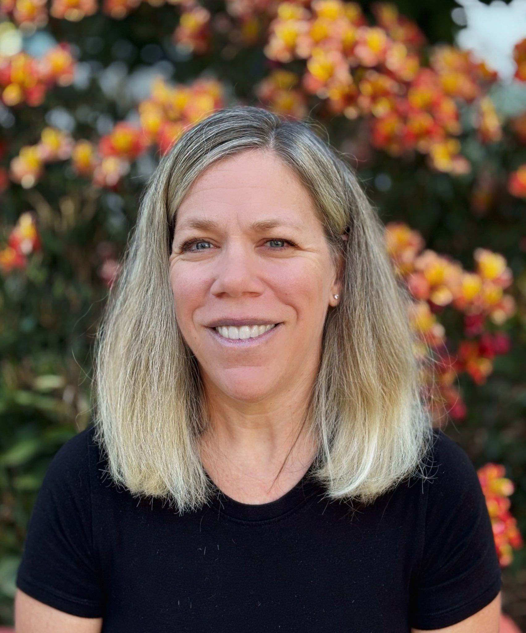 Woman with blonde hair smiling in front of orange and yellow flowers, wearing a black shirt.
