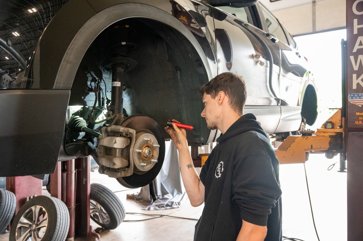 Mechanic examines car brakes on a lift. He wears a black hoodie, holds a light, and is in a shop.