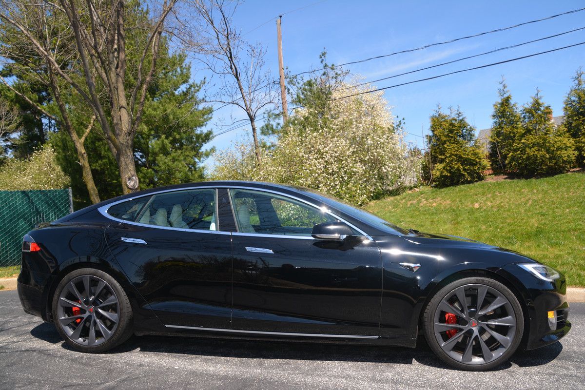 Black Tesla sedan parked on a paved road, with greenery and a blue sky in the background.