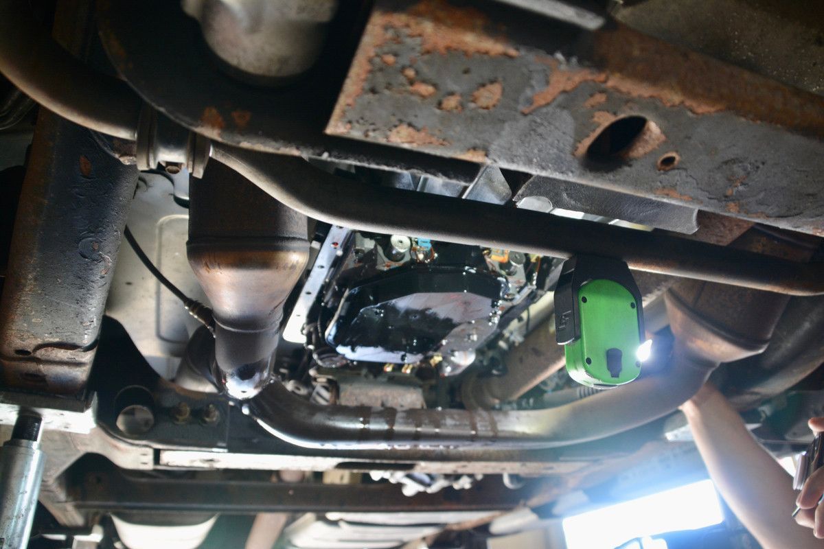 Underneath a car, a person examines a black, oily transmission pan with a flashlight; the setting is a garage.