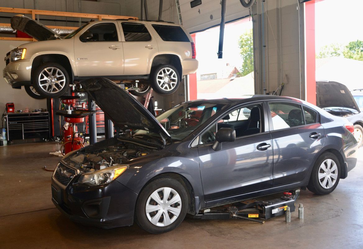 A gray sedan with an open hood in a car repair shop, with a tan SUV elevated above.