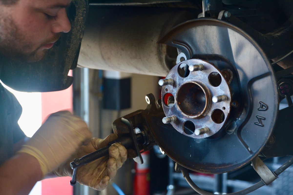 Mechanic repairing a car's brake system, wearing gloves, in a garage setting.