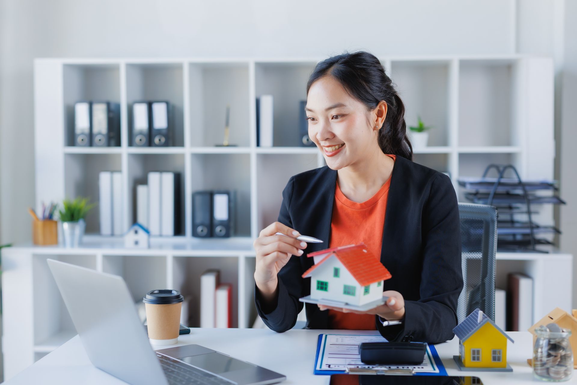 Woman holding house model, pointing at it while on a laptop video call in an office.