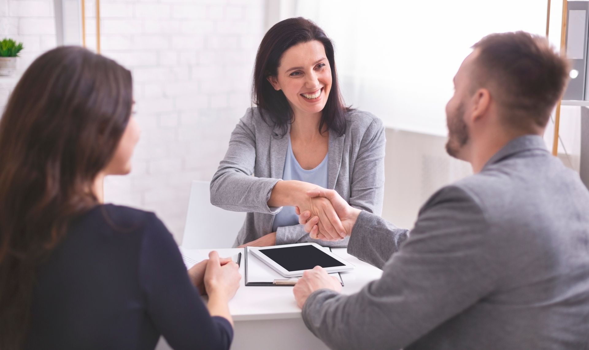 Three people in business attire shake hands around a white table. Smiling, they are indoors.