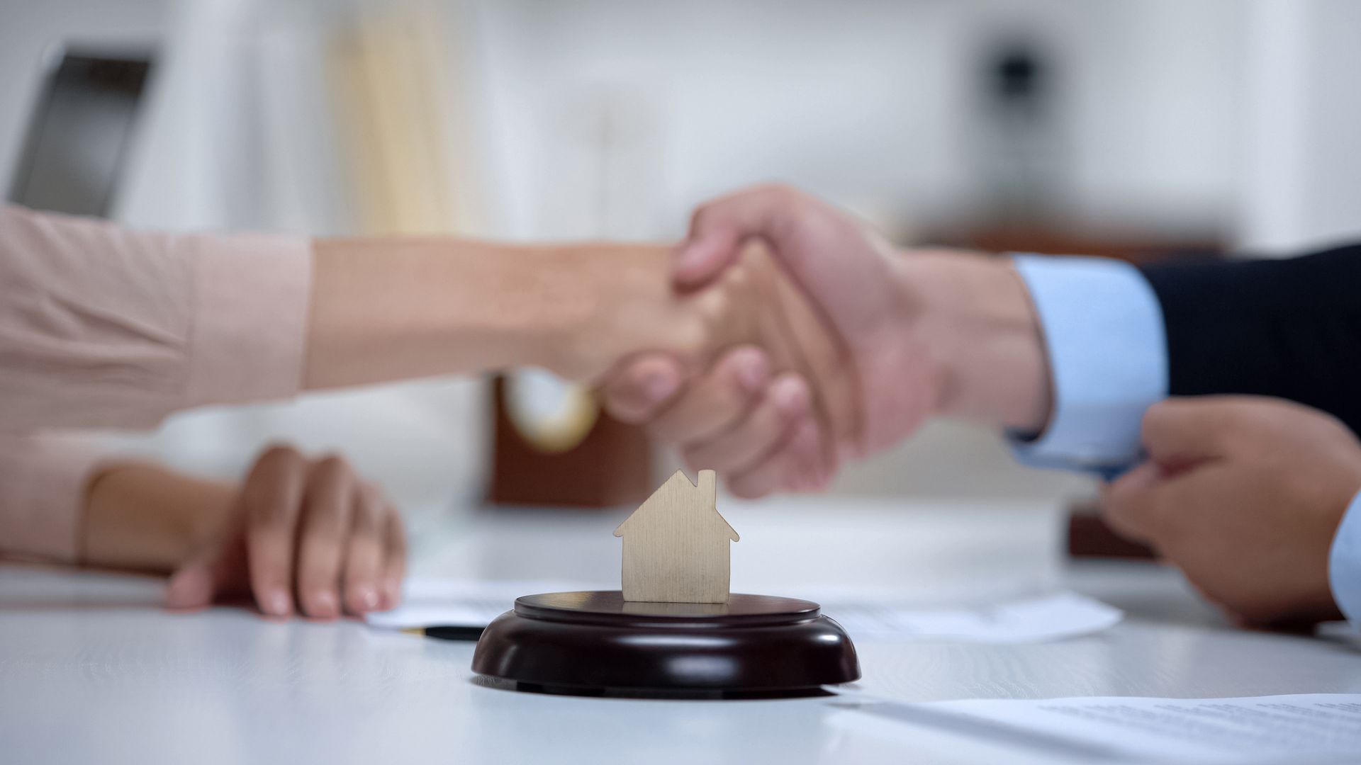 Two people shake hands over a wooden house model on a table, possibly closing a real estate deal.