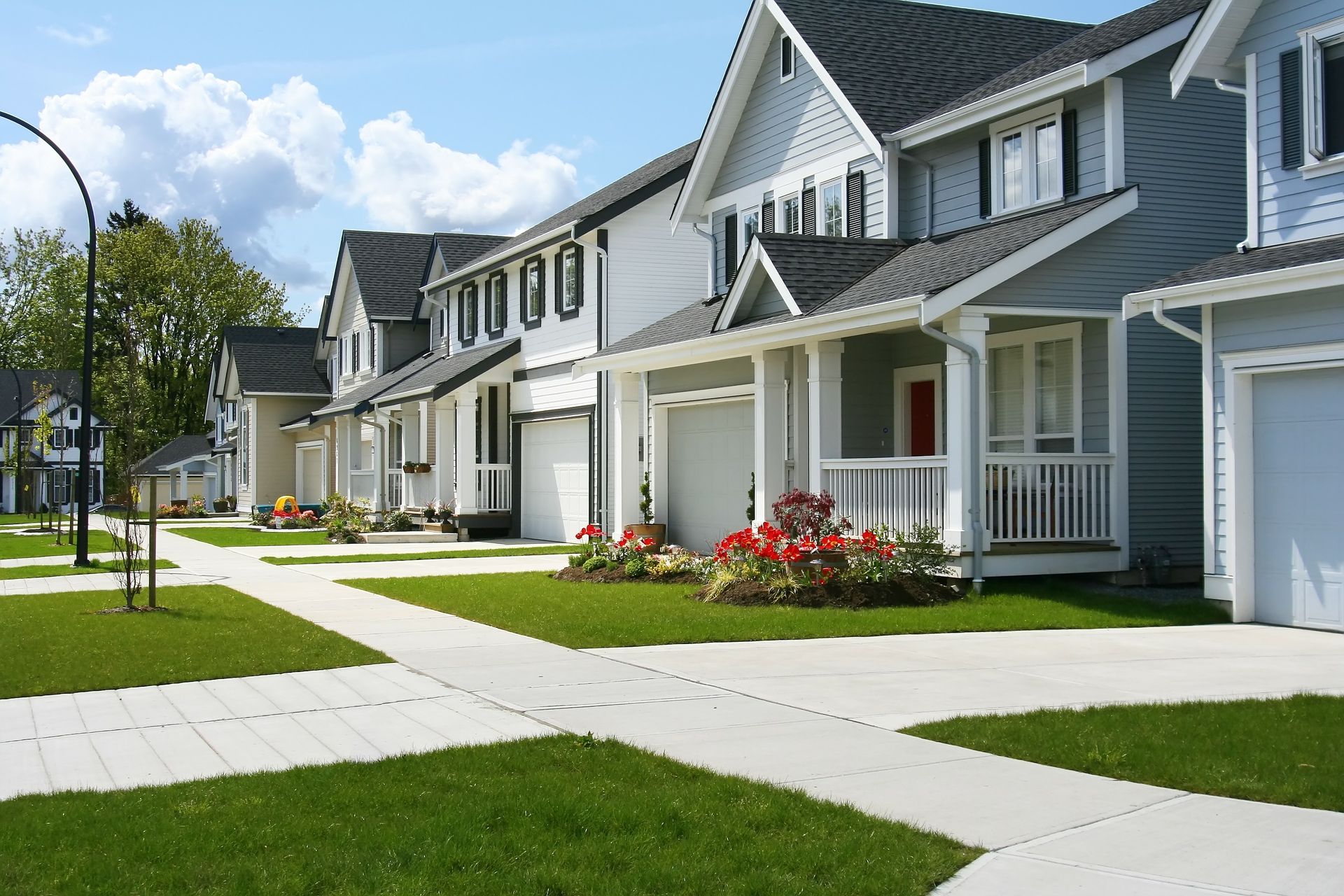 Row of suburban houses with garages, green lawns, and sidewalks under a blue sky with fluffy clouds.