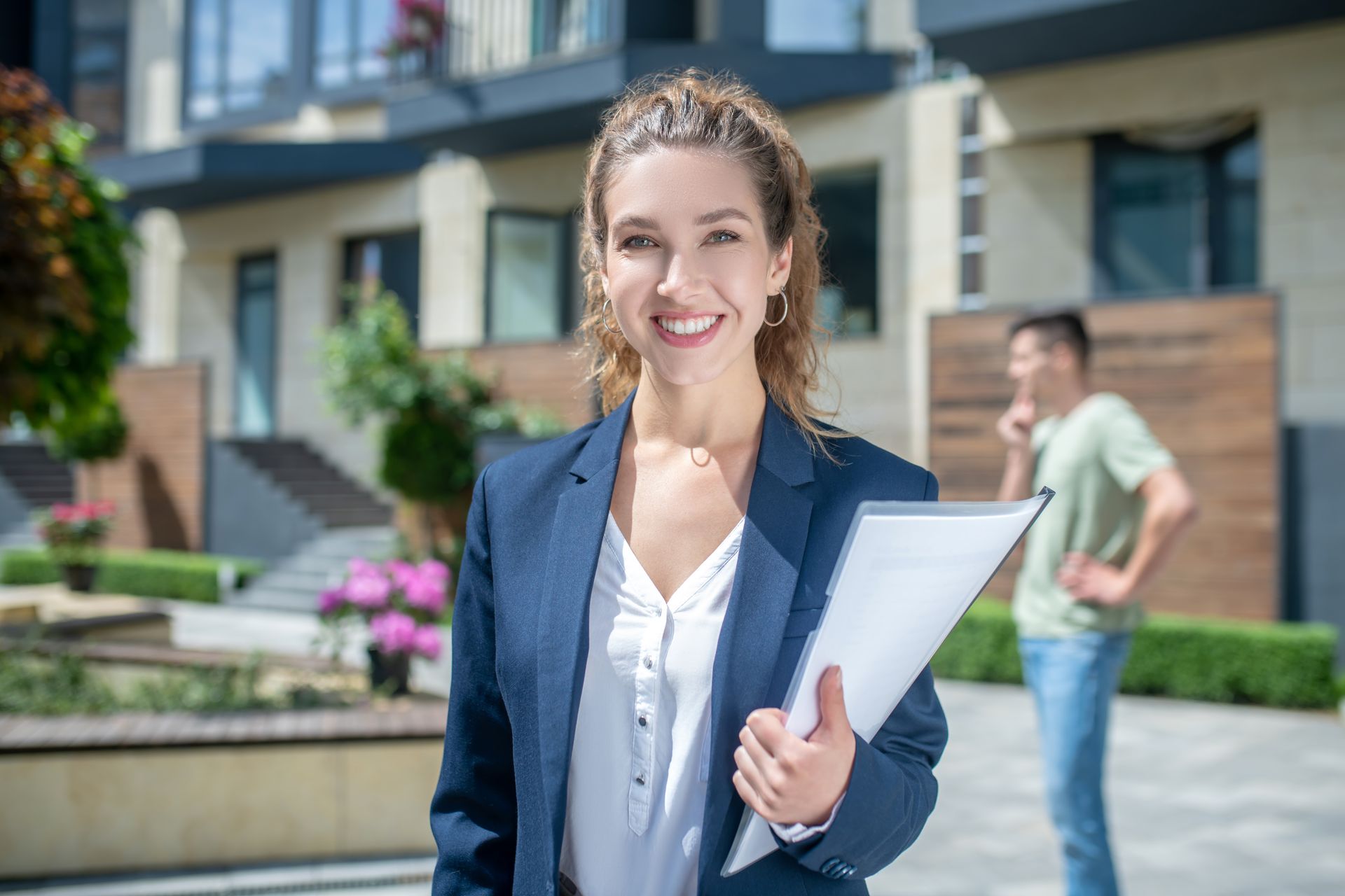 Woman in a blazer smiles, holding a file, standing in front of a building with a man in the background.