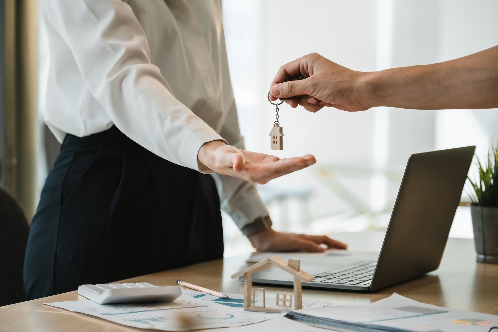 Hand holding house key over outstretched hand. Desk with laptop, documents, and house model.