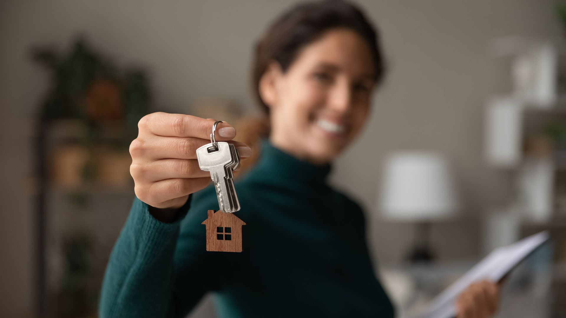 Woman holding up keys with house-shaped keychain, smiling indoors.