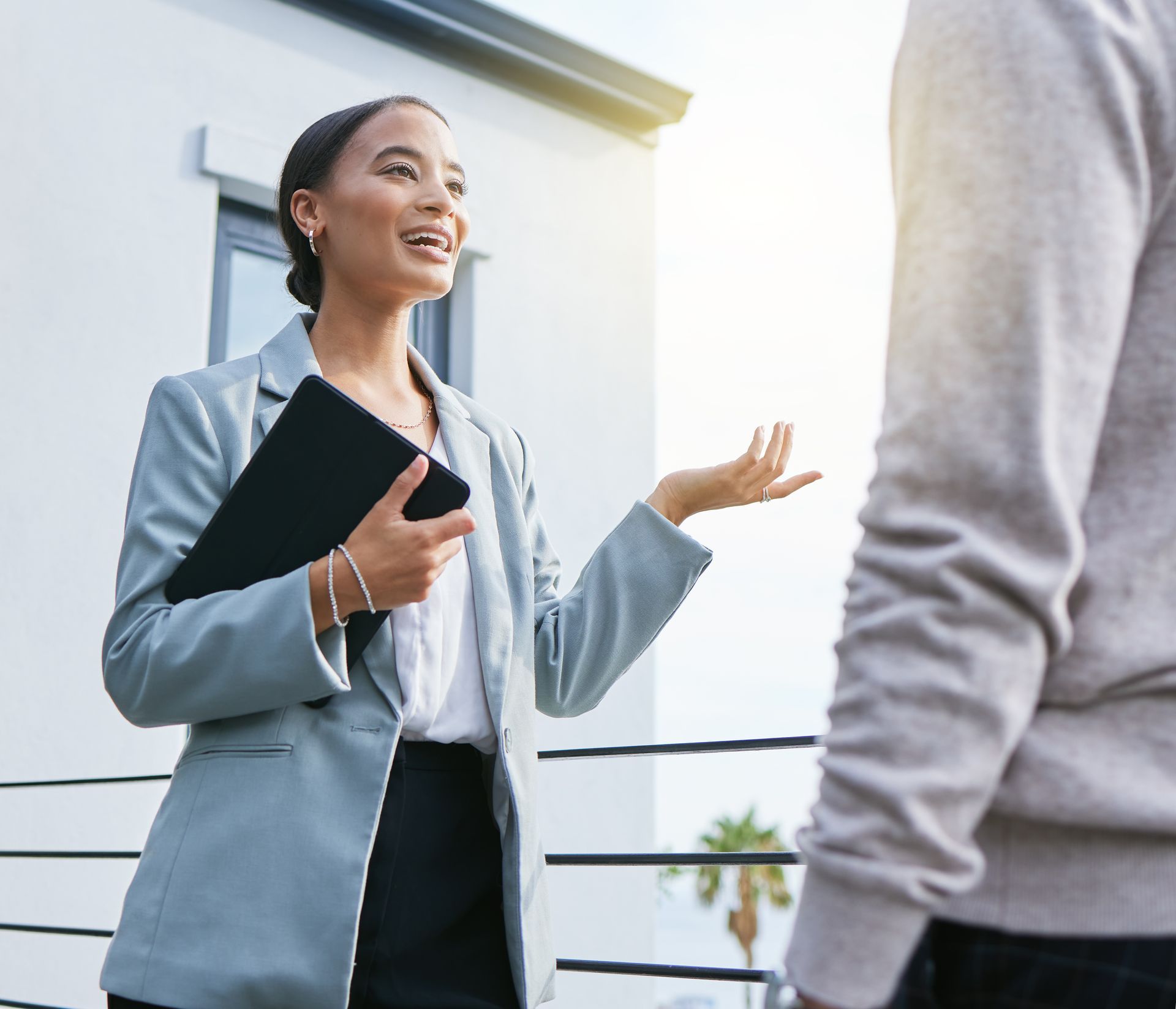 Woman in a blazer holds a tablet, gestures, and speaks to another person outside a building.