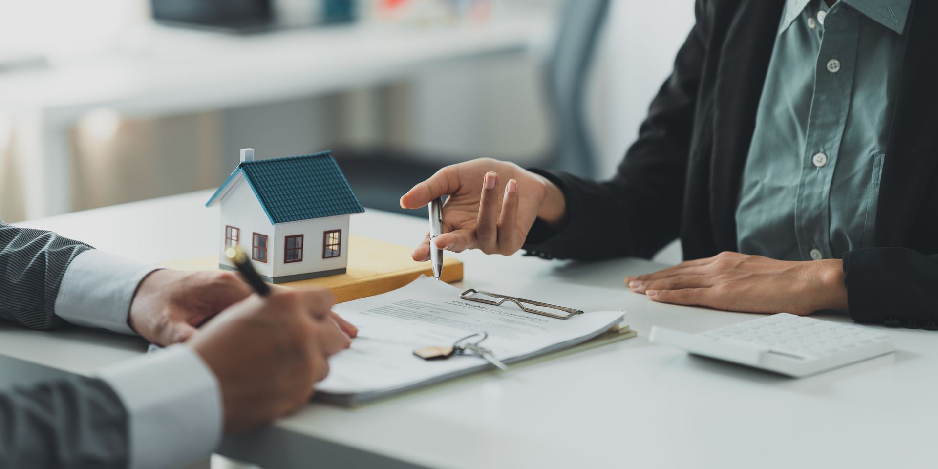 Person signing paperwork with another person at a table, a model house and keys present.