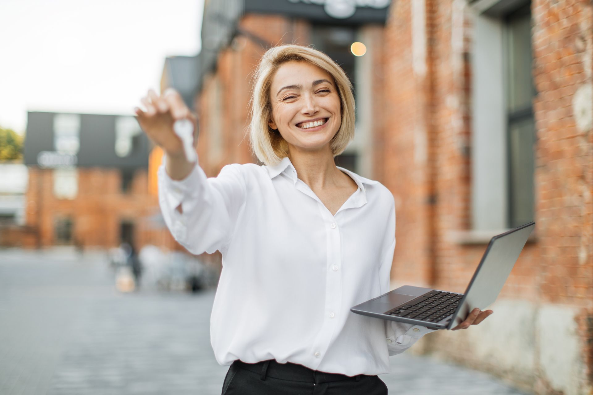 Woman with keys and laptop smiles, standing outdoors in front of a brick building.