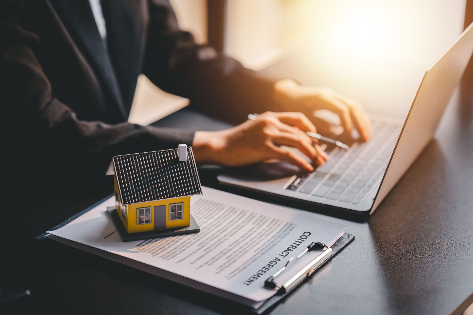 Person typing on laptop, small house model and contract on desk, lit by sunlight.