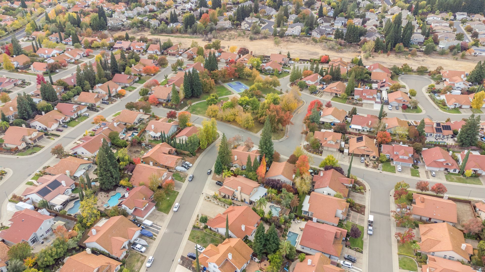 Aerial view of a suburban neighborhood with rows of houses, red roofs, trees, and roads.