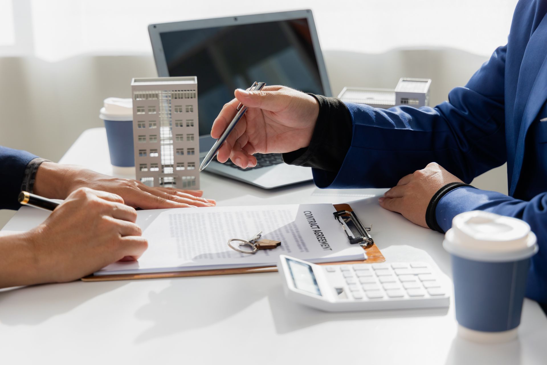 Two people reviewing documents, models of buildings, laptop, calculator, coffee cups on a table.