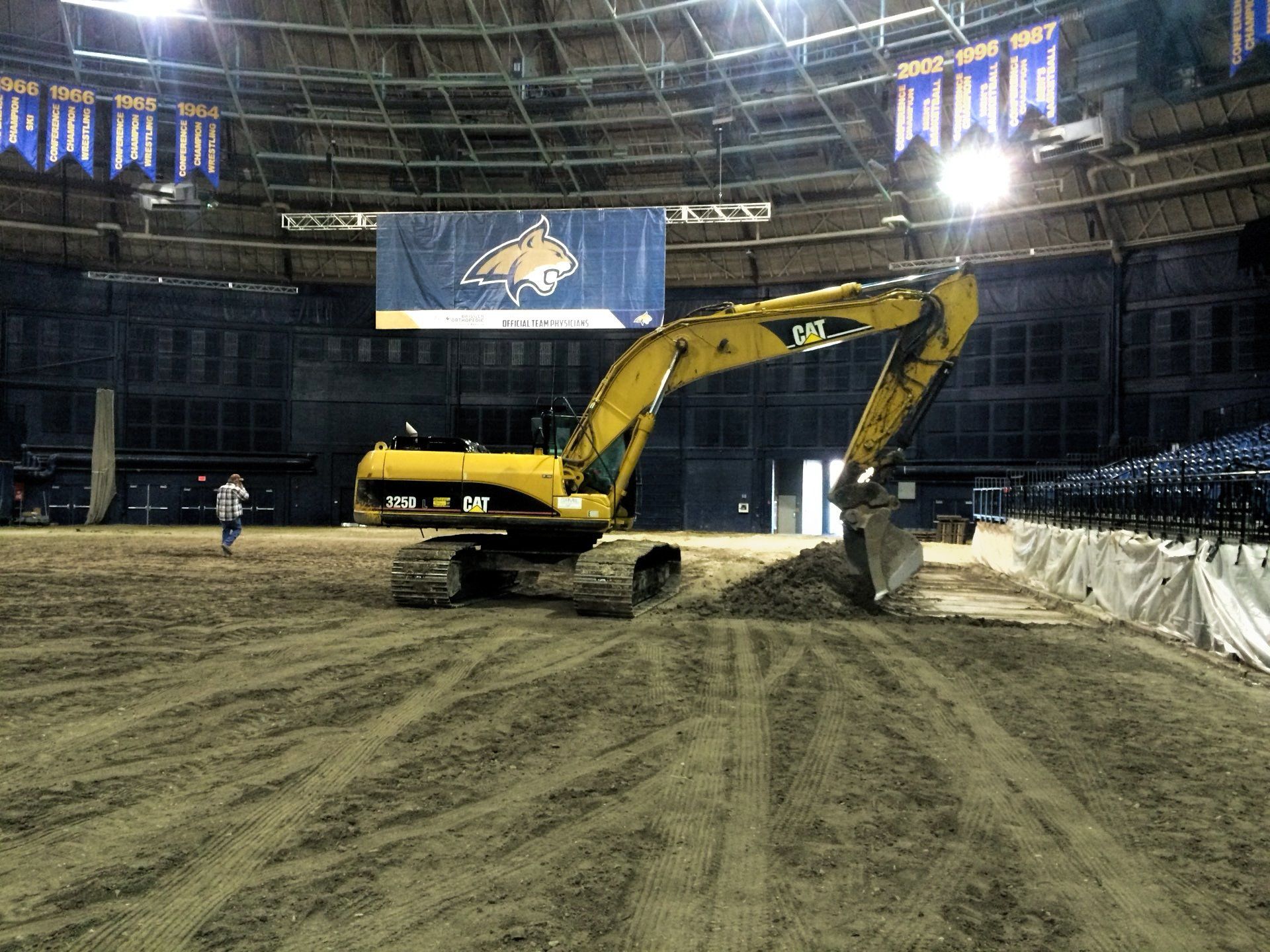 A large yellow excavator is moving dirt in a stadium.