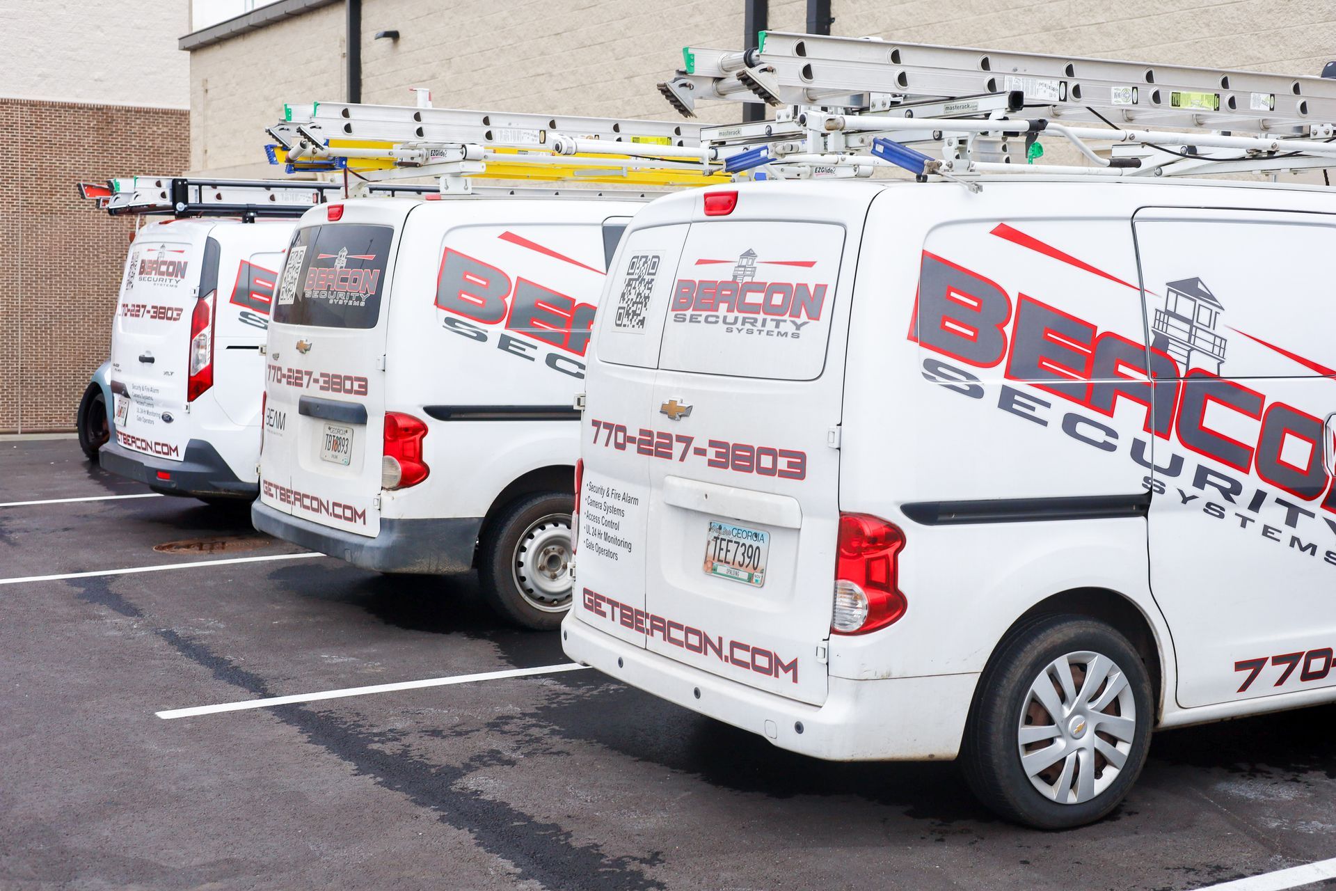 Three white Beacon Security Systems vans parked in a row in a paved parking lot.
