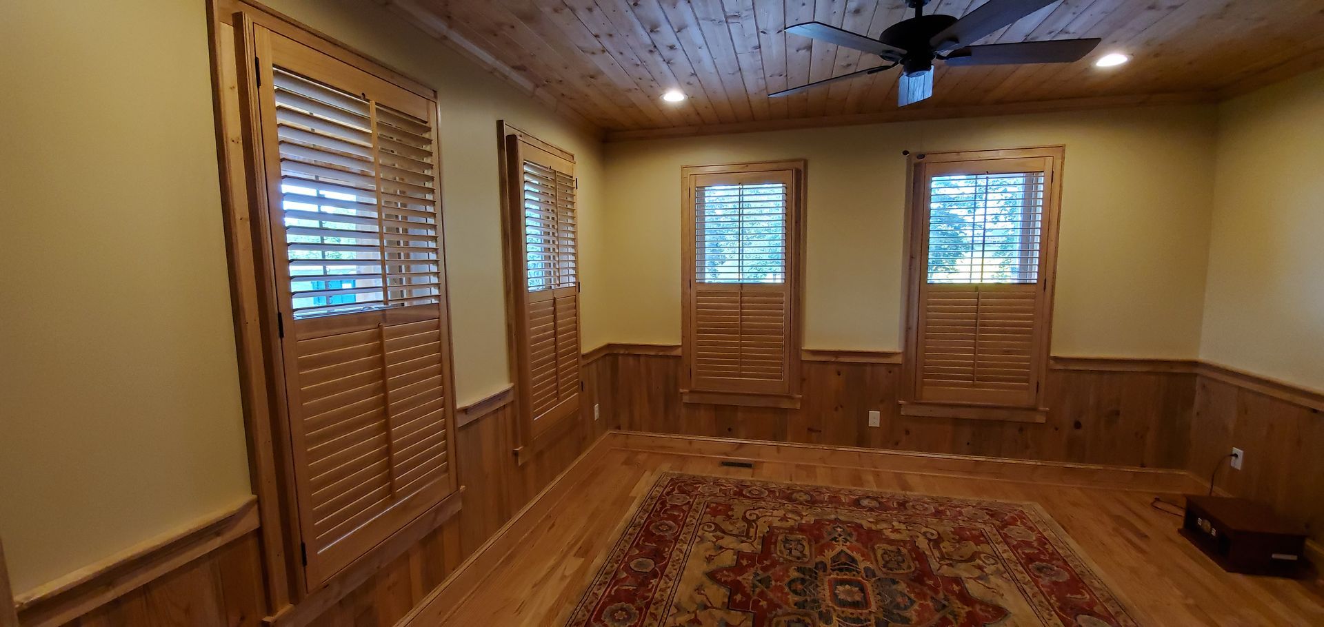 Empty wood-paneled room with patterned rug, windows, and a ceiling fan.