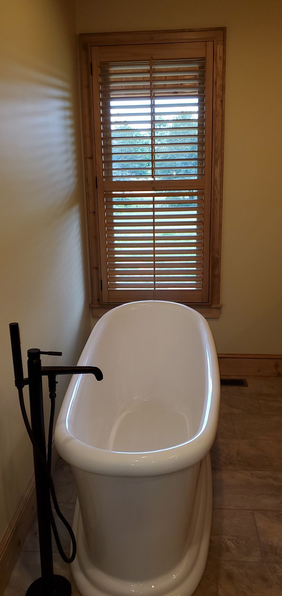 Bathroom with white freestanding bathtub, black faucet, and wood-framed window with blinds.