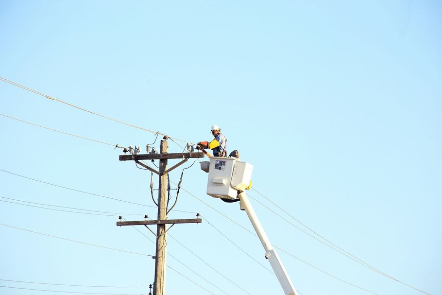 A utility worker in a white bucket truck performs repairs on power lines attached to a wooden utility pole against a sky.