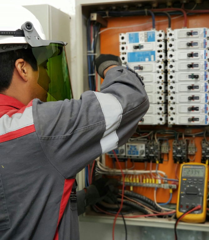 An electrician in protective gear tests components in an open electrical panel using a yellow multimeter.