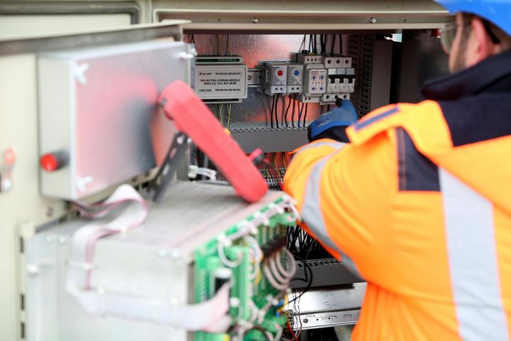 A technician in high-visibility orange workwear inspects electrical wiring inside a control panel.