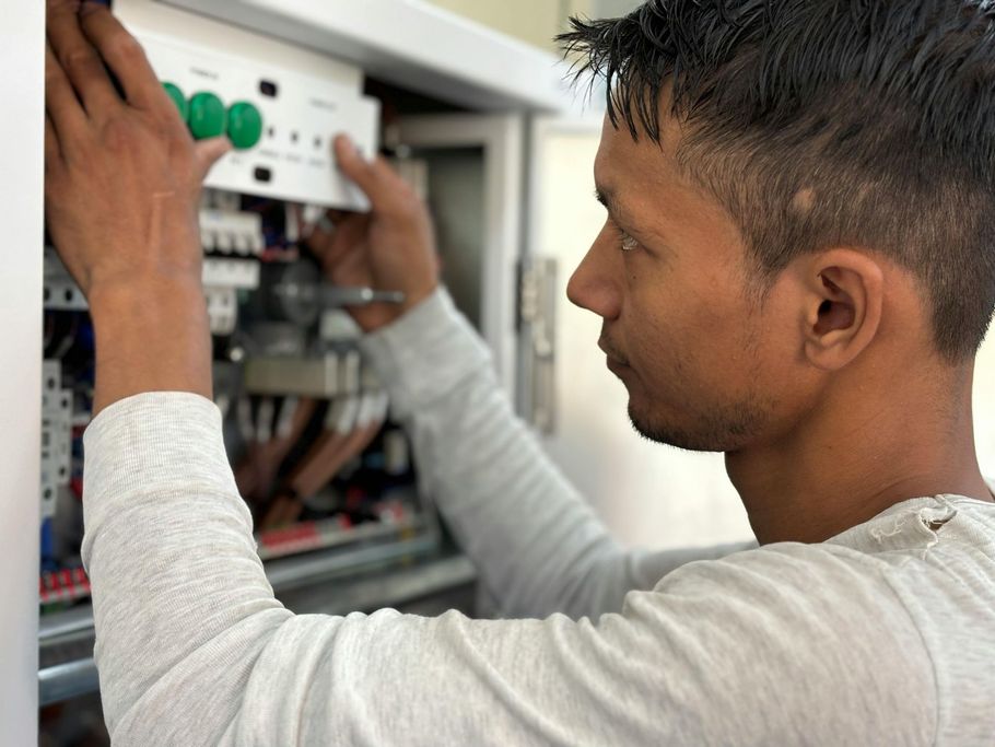 A person in a light gray long-sleeved shirt installs or repairs a white electrical control panel.