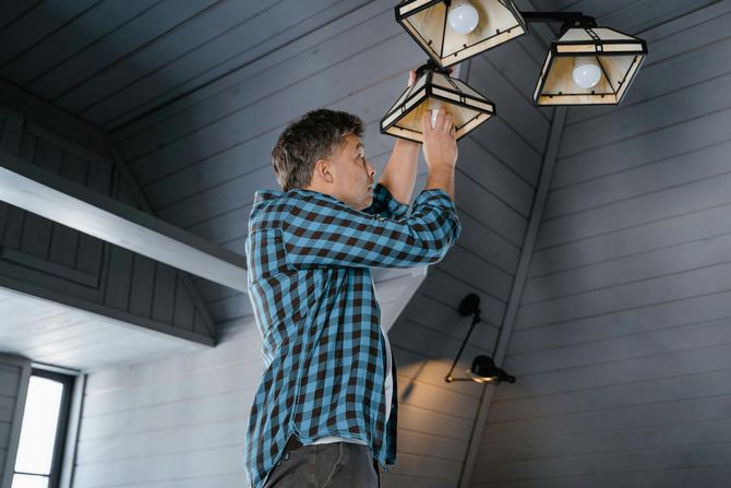 A person in a blue plaid shirt stands on a ladder, reaching up to change a lightbulb in a wooden ceiling fixture.