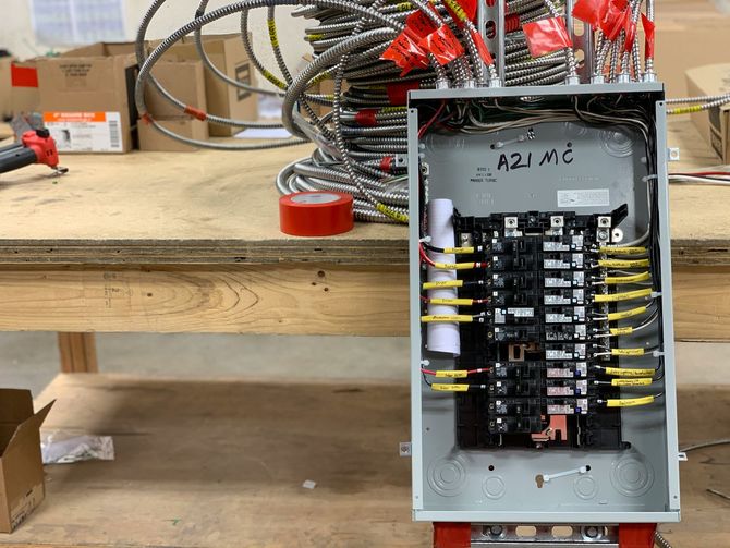 An open electrical load center with wired circuit breakers mounted on a workbench beside coiled armored cables.