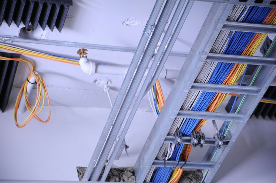 A low-angle view of a ceiling with a metal cable tray holding bundles of multicolored data cables and a fire sprinkler.