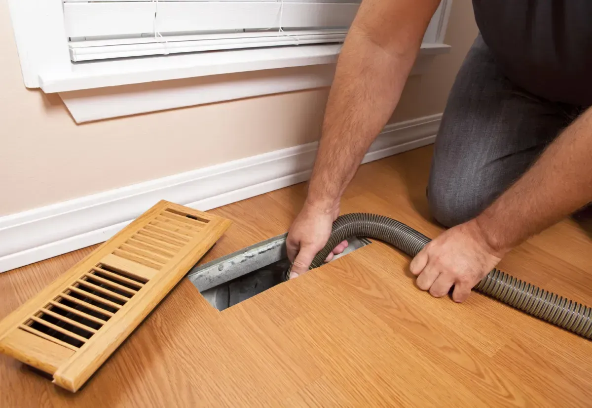 A man is using a vacuum cleaner to clean a vent on the floor.