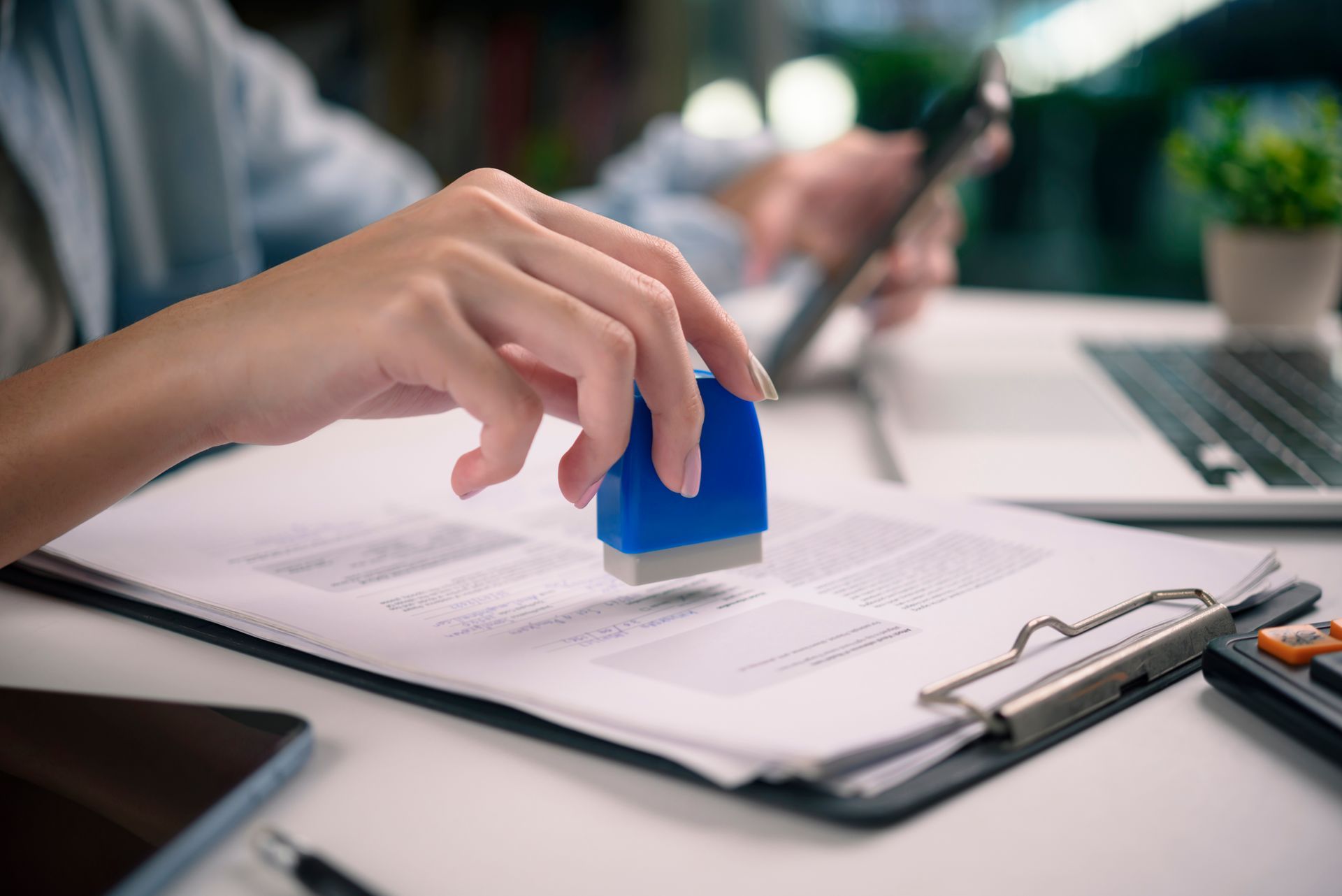 Person stamping a document with a blue ink stamp; workspace with laptop, calculator, and tablet.