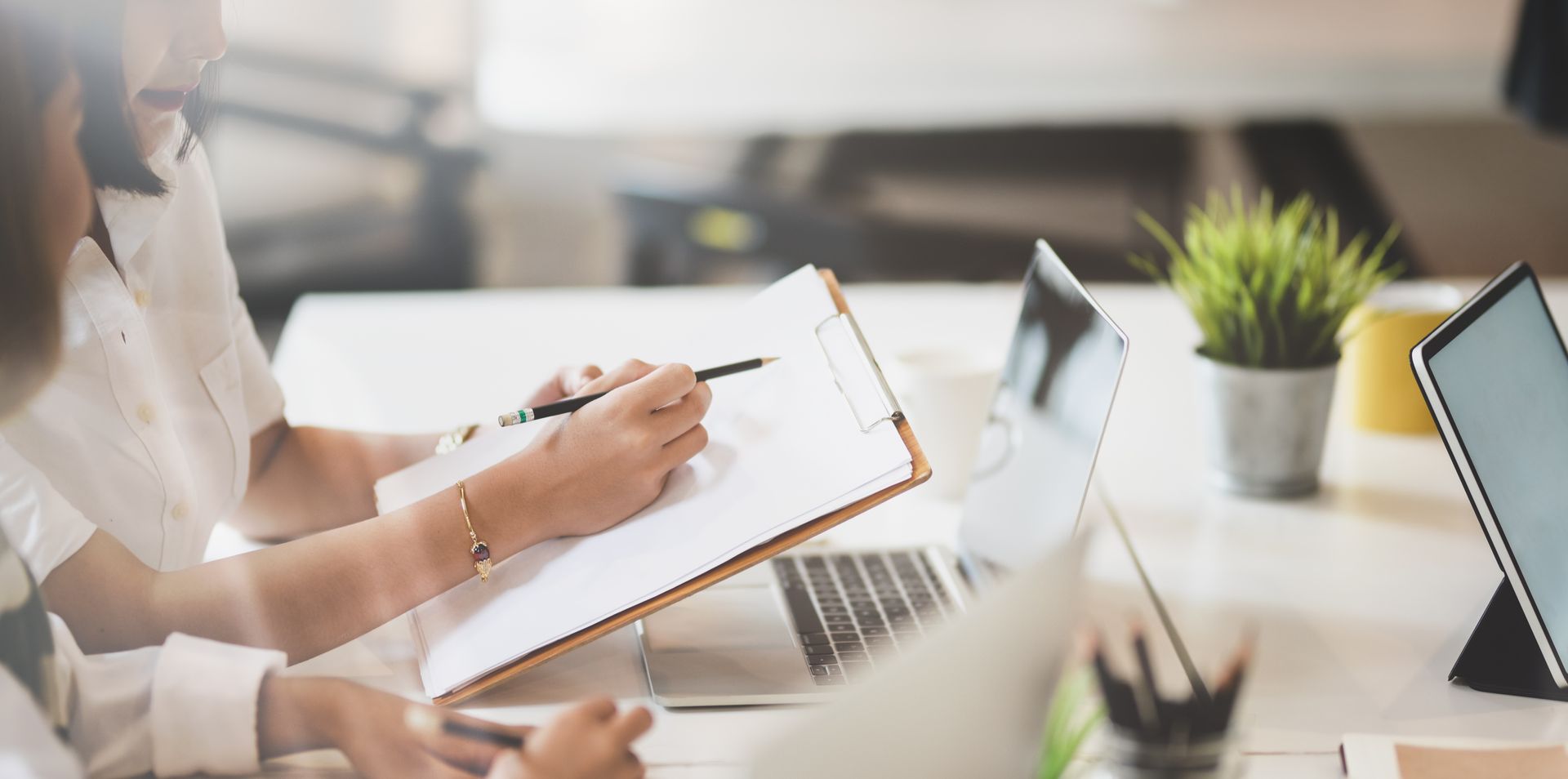 Two people working at a table, one writing on a clipboard, laptop and tablet visible.