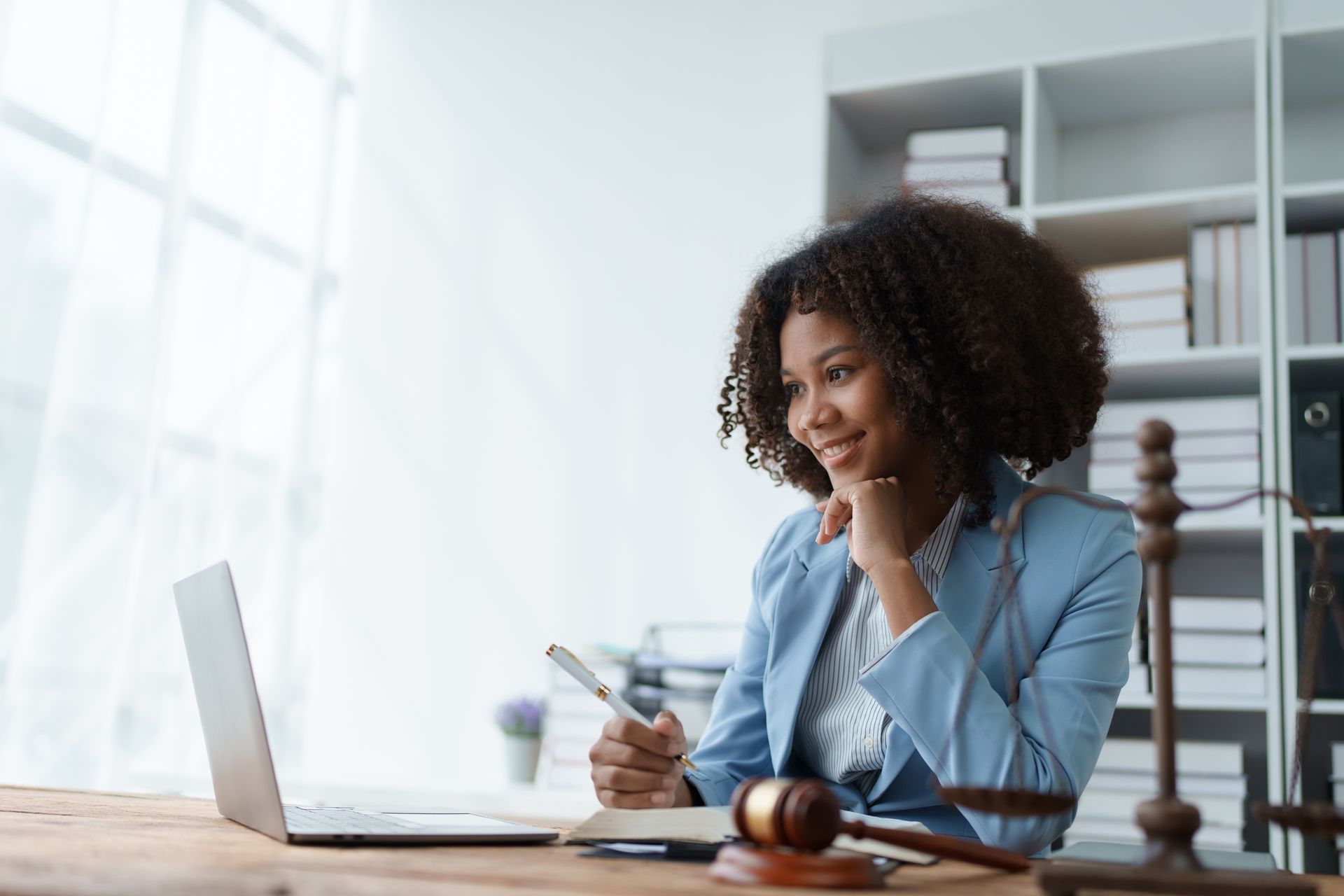 Woman in blue blazer, smiling, at a desk with a laptop, gavel, and notebook. Office setting.