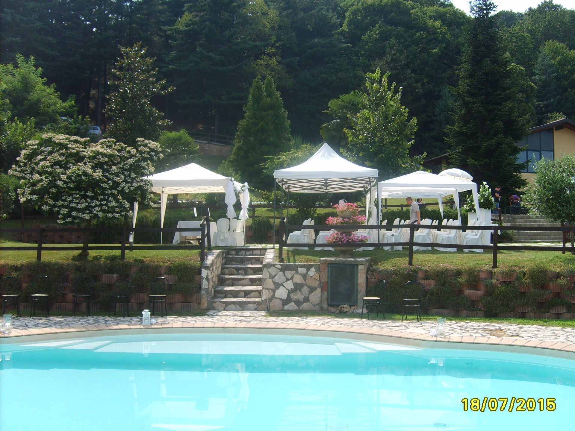 Piscina con tre gazebo bianchi su una collina erbosa; alberi sullo sfondo.