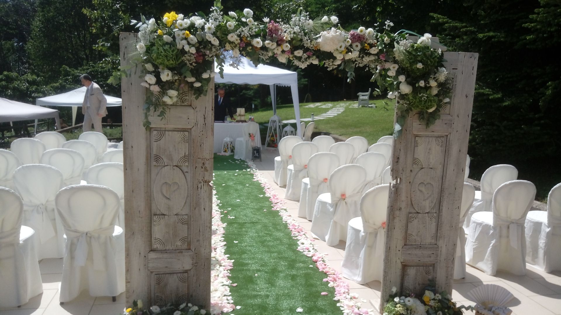 Cerimonia nuziale all'aperto con arco floreale, navata con tappeto verde e file di sedie bianche.