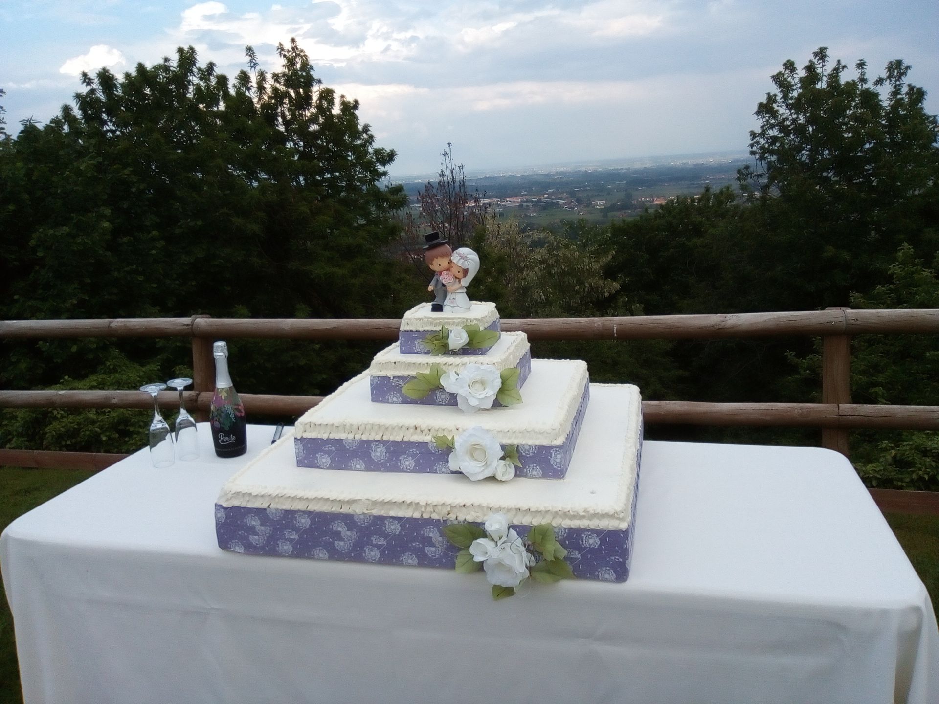 Torta nuziale a tre piani su un tavolo all'aperto; piccole figure in cima, che si affacciano sul paesaggio.