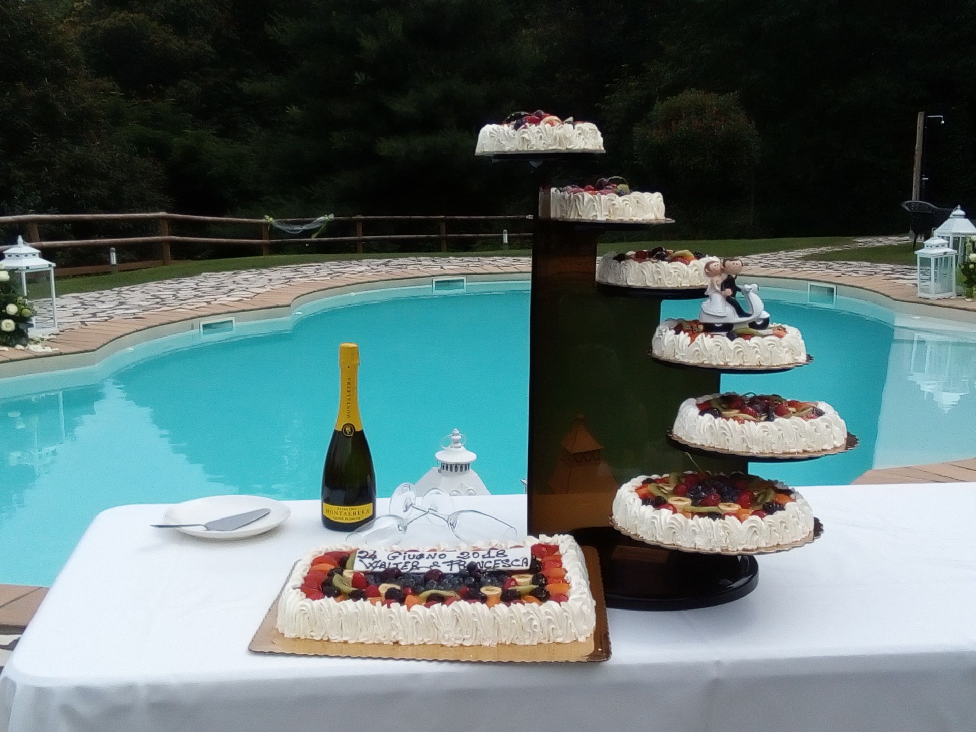 Torte nuziali a bordo piscina; torte rotonde a più piani e una torta rettangolare con guarnizione di frutta.