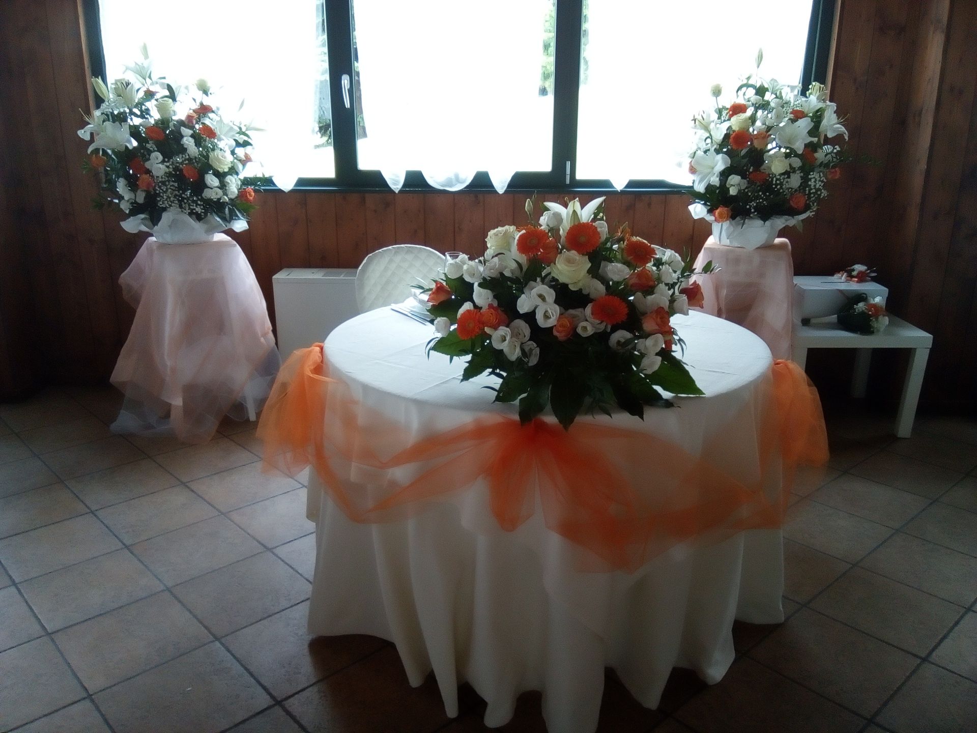 Tavolo rotondo con tovaglia bianca, tulle arancione e centrotavola floreale; ai lati composizioni floreali su piedistalli drappeggiati.