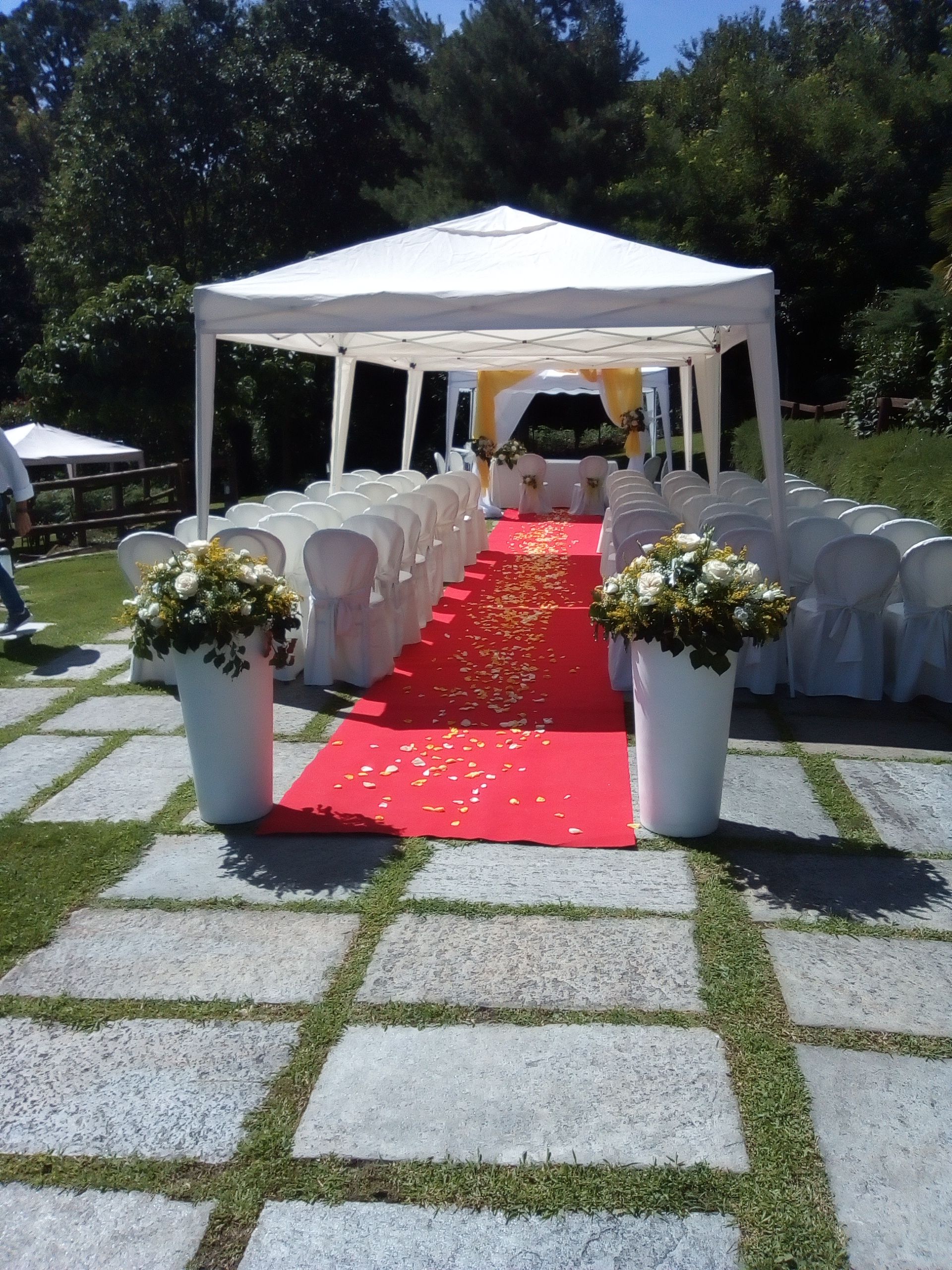 Allestimento della cerimonia nuziale: corridoio con tappeto rosso, tenda bianca, composizioni floreali, file di sedie, allestimento esterno.